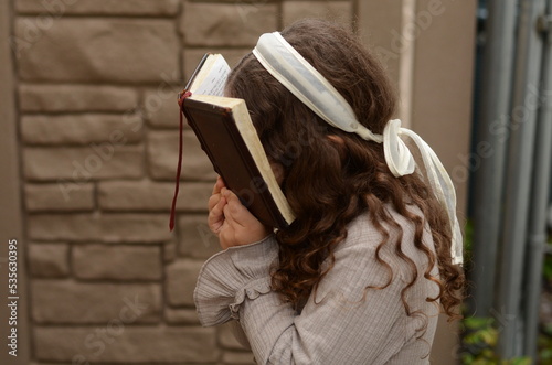 Little pretty Jewish girl with curly hair, wearing a dress and a white ribbon headband praying from a prayer book, tehillim, siddur outdoors photo