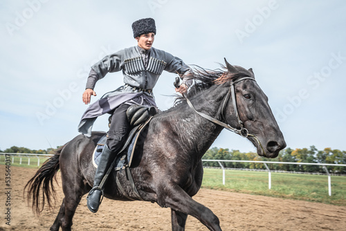 Cossack man in a national costume is galloping rapidly on a black horse around the hippodrome photo