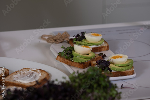 white plate with delicious toast with salted salmon, avocado, egg and greens on white kitchen table with blurred kitchen background