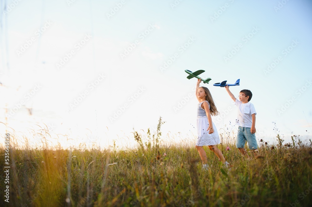 Running boy and girl holding two green and blue airplanes toy in the field during summer sunny day Running boy and girl holding two green and blue airplanes toy in the field during summer sunny day