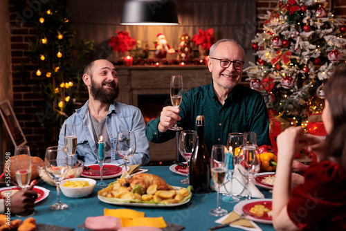 Smiling man proposing christmas toast at festive dinner, holding glass with sparkling wine at xmas home feast. Family celebrating winter holiday, drinking at new year party