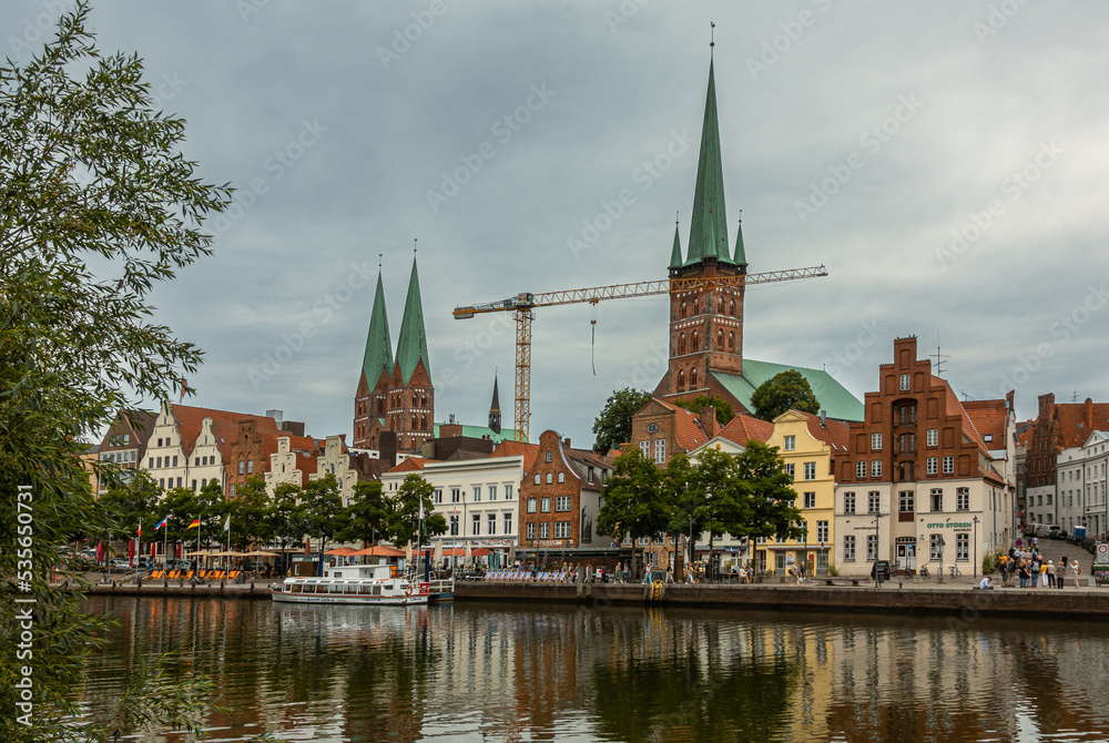 Germany, Lubeck - July 13, 2022: 2 churches seen from over Trave river. 1 tower S. Petri, Peter, and 2 towers St. Marien, Mary, under gray cloudscape. historic architecture on quay