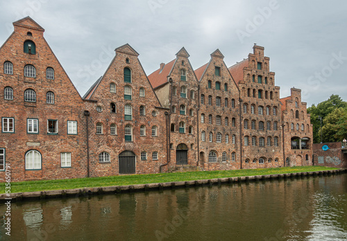 Germany, Lubeck - July 13, 2022: Row of red-brown historic salt storage warehouses along Trave river under blueish cloudscape. Green lawn on quay.