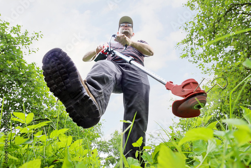 View from bottom up from ground, leg comes close-up, person walks along grass, trimer is in hands