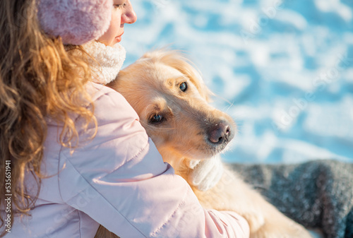 Portrait of young beautiful woman in pink headphones with curly hair sitting and hugging her golden retriever dog. Happiness and friendship. pet and woman. Winter landscape on the laying of the river 