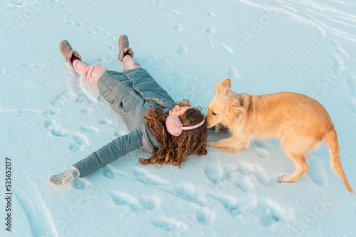Long curly young girl in pink winter earmuffs and white mittens plays with her dog on winter cold ice river covered with snow. Snowing winter concept.
