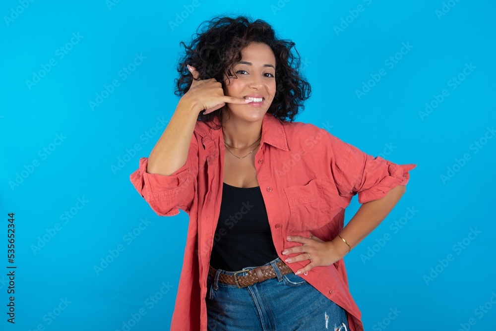 young beautiful woman with curly short hair wearing red overshirt over blue wall imitates telephone conversation, makes phone call gesture with hands, has confident expression. Call me! young beautiful woman with curly short hair wearing red overshirt over blue wall imitates telephone conversation, makes phone call gesture with hands, has confident expression. Call me!
