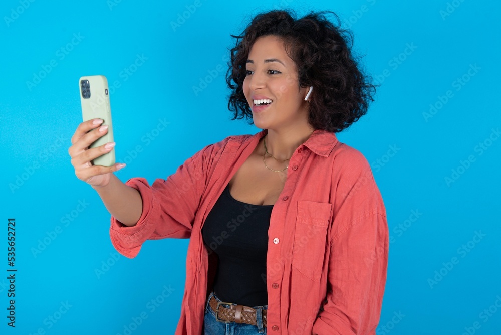 Isolated shot of pleased cheerful young beautiful woman with curly short hair wearing red overshirt over blue wall, makes selfie with mobile phone. People, technology and leisure concept Isolated shot of pleased cheerful young beautiful woman with curly short hair wearing red overshirt over blue wall, makes selfie with mobile phone. People, technology and leisure concept