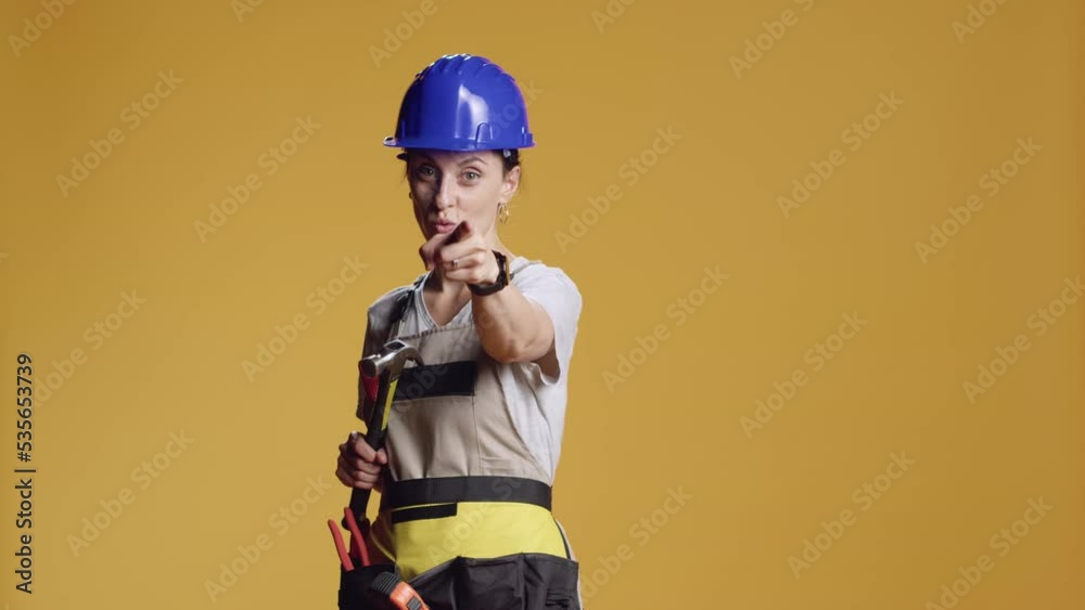 Portrait of engineer holding hammer and pointing at camera, being aggressive with breaking tool. Handywoman with hardhat using index finger to point in studio, holding sledgehammer. Portrait of engineer holding hammer and pointing at camera, being aggressive with breaking tool. Handywoman with hardhat using index finger to point in studio, holding sledgehammer.