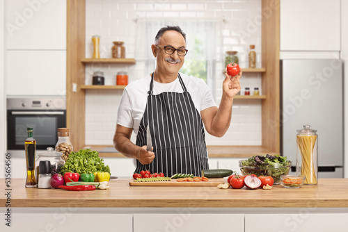 Mature man behind a counter holding a knife and a tomato in a kitchen