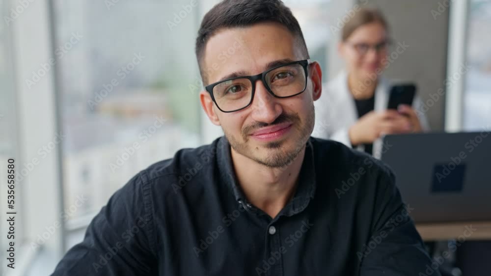 Positive smiling male specialist working in the office. Portrait of an average office employee working at laptop. Female speaking on the phone at backdrop. Positive smiling male specialist working in the office. Portrait of an average office employee working at laptop. Female speaking on the phone at backdrop.