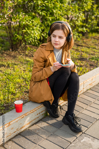A young teenage girl sits on the street with a smartphone in her hand, headphones, listens to music. High quality photo