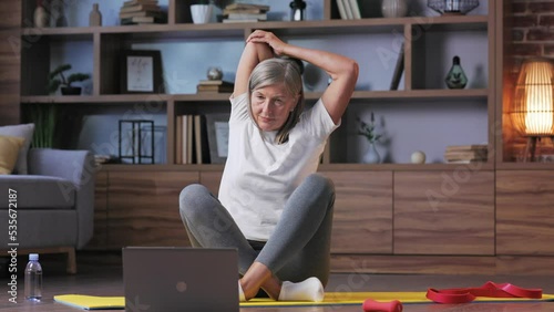Gray-haired happy mature woman doing gymnastics and yoga at home online with laptop. Active and healthy lifestyle for pensioners. Fitness lessons online, recreation, well-being of the elderly.