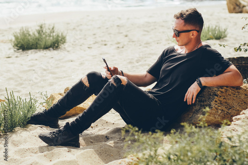 A fit white man in a black T-shirt and jeans sits on the ocean leaning on a stone and looks at the smartphone screen.