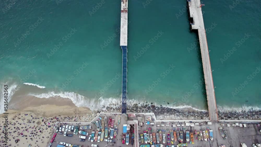 imagen aerea cenital desde dron de una playa y un par de muelles en donde se ven botes de colores y el mar de color turquesa con olas