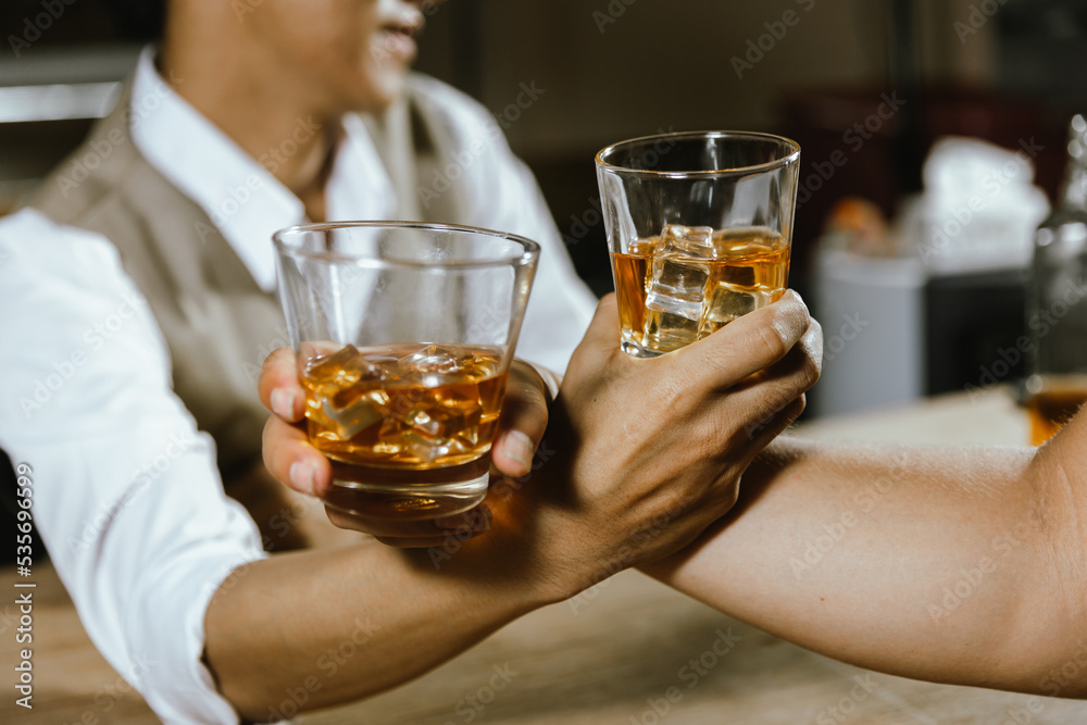 A close-up shot of two men clinking whiskey glasses together while at the bar counter in a pub.