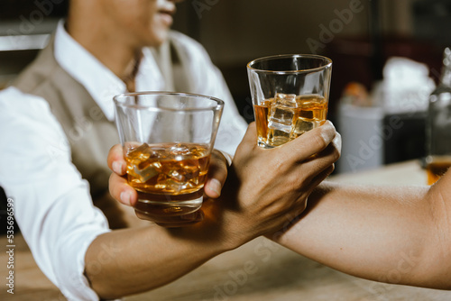 A close-up shot of two men clinking whiskey glasses together while at the bar counter in a pub.