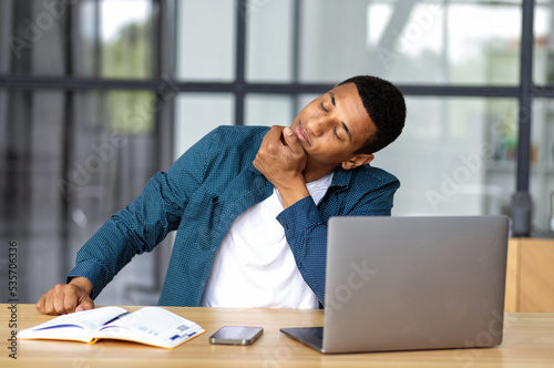 Tired African American businessman man working, sitting at the workplace in the office stretching his neck, doing a warm-up at desk