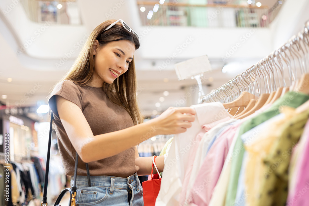 Happiness young Beautiful woman with shopping bag choosing dress at row of clothes fashion store. Trendy female at retail mall. Women shopping lifestyle fashion Happiness young Beautiful woman with shopping bag choosing dress at row of clothes fashion store. Trendy female at retail mall. Women shopping lifestyle fashion