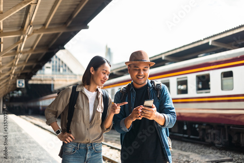 Couple Young asian traveler tourist by train Choose a trip and booking train queue by smartphone. Two Backpacker at platform train station. Happy traveller at railway on vacation holiday weekend. photo