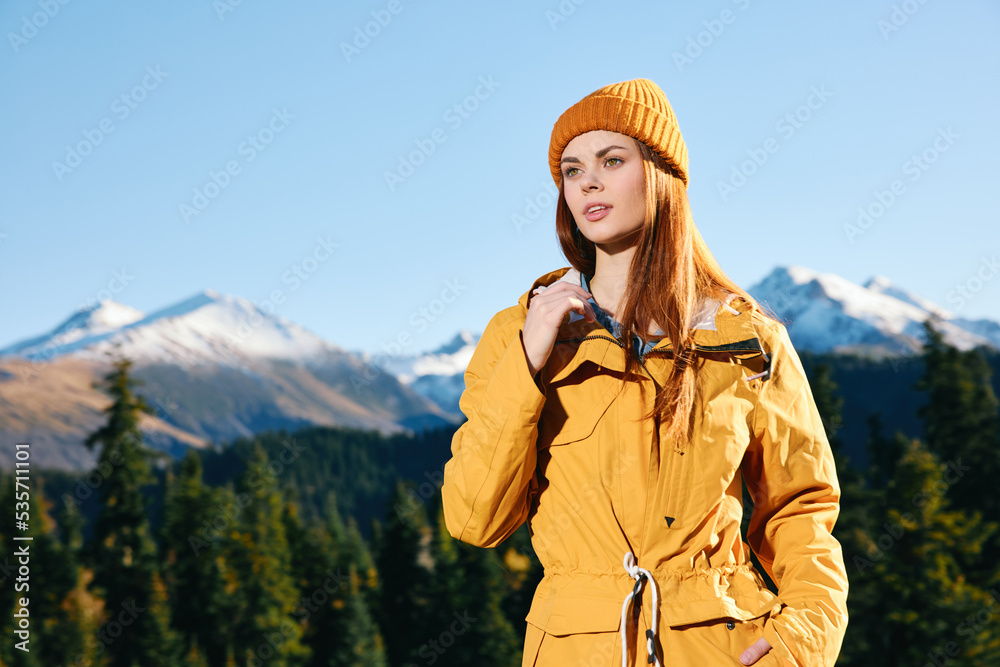 Woman with red hair hiker in yellow raincoat and cap standing on a mountain overlooking snowy mountains traveling in winter and hiking in the mountains in the wilderness at sunset