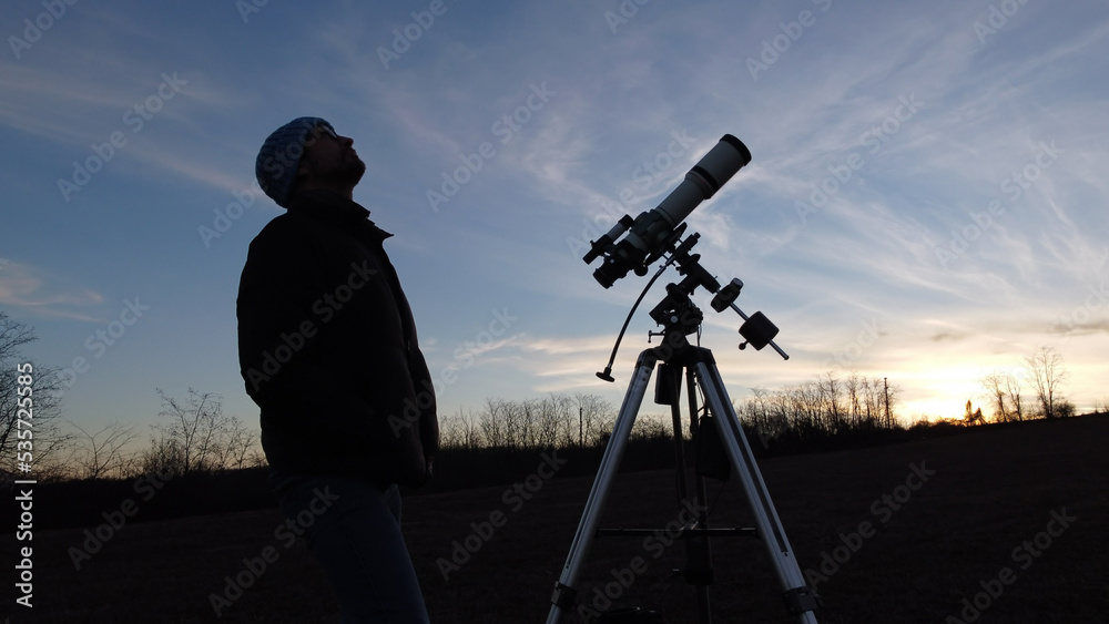 Silhouette of a man, telescope and countryside under evening skies.