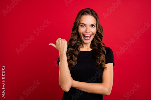 Photo of adorable excited young woman wear black t-shirt pointing thumb empty space smiling isolated red color background © deagreez