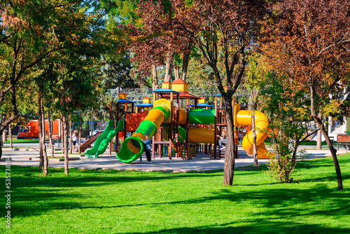 bright sunny day in autumn city park, children play on the playground, green lawn and yellow leaves, street
