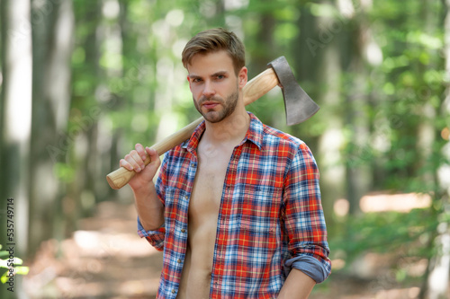 Lumbersexual guy in lumberjack shirt holding axe on shoulder forest background