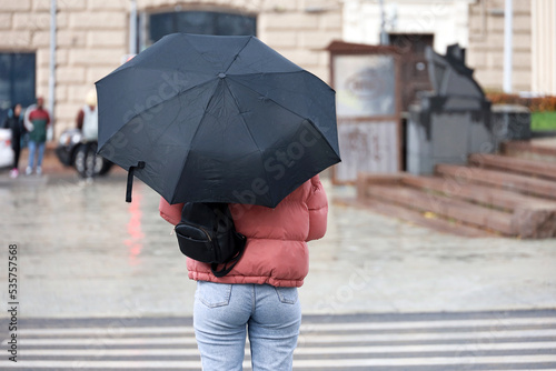 Rain in city, slim woman with umbrella wearing jeans and jacket standing on а street on crosswalk background. Rainy weather in autumn