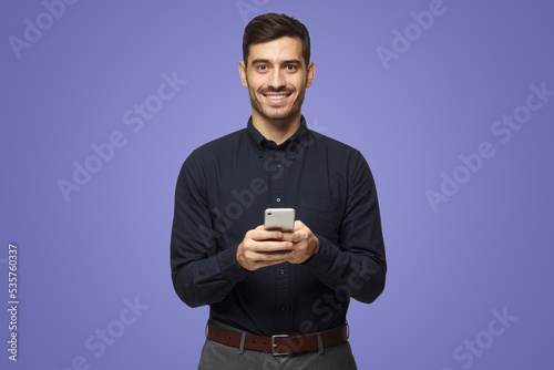 Young business man standing isolated on purple holding phone, looking at camera and smiling nicely © Damir Khabirov