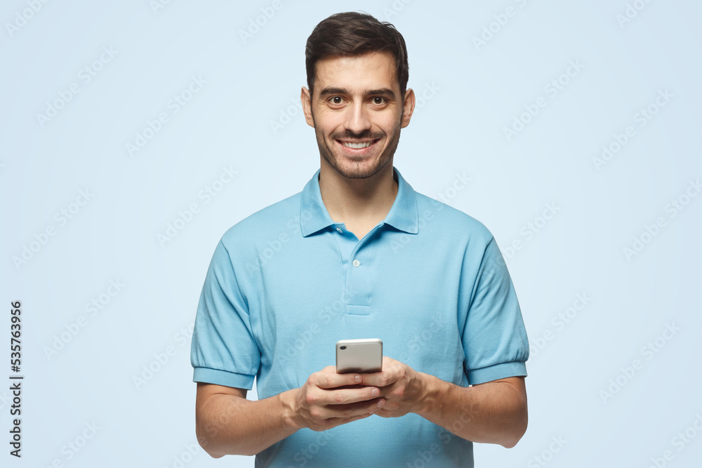 Smiling man standing on blue background, looking at camera, holding phone with both hands Smiling man standing on blue background, looking at camera, holding phone with both hands