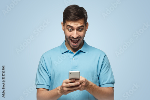 Excited man looking at his phone with surprise, isolated on blue background © Damir Khabirov