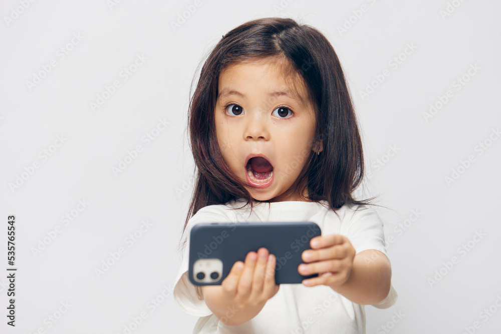 funny, cheerful, emotional little girl in a white t-shirt stands holding a trendy smartphone in her hand and looks at the camera with her mouth wide open. funny, cheerful, emotional little girl in a white t-shirt stands holding a trendy smartphone in her hand and looks at the camera with her mouth wide open.