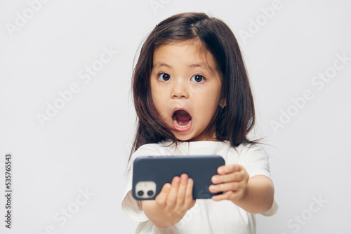 funny, cheerful, emotional little girl in a white t-shirt stands holding a trendy smartphone in her hand and looks at the camera with her mouth wide open.