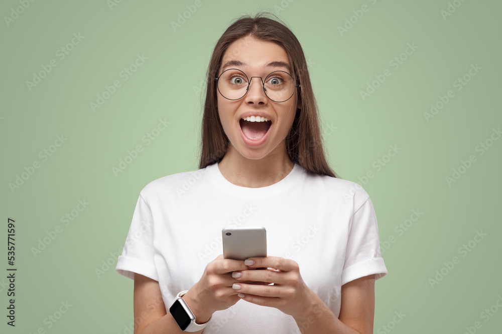 Close-up of girl in white t-shirt, holding phone, looking surprised and shocked, isolated on green Close-up of girl in white t-shirt, holding phone, looking surprised and shocked, isolated on green