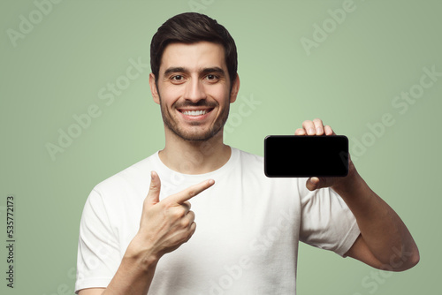 Happy man pointing with finger at blank smartphone, smiling at camera, isolated on green background