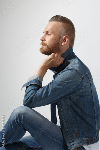 Stylish attractive white man with a beard in jeans sits isolated on a light gray background. © ksi