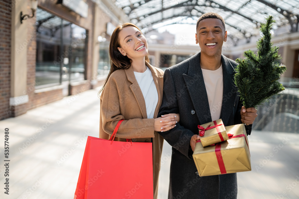 cheerful interracial couple with shopping bags and gift boxes with small christmas tree looking at camera near mall. cheerful interracial couple with shopping bags and gift boxes with small christmas tree looking at camera near mall.