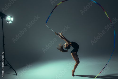 Gymnast child girl performs gymnastic exercises with ribbon in her hand lit by spotlight.