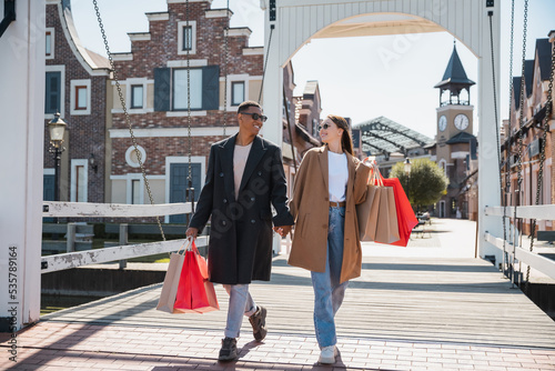 full length of cheerful and trendy multiethnic couple holding hands while walking on city bridge with shopping bags