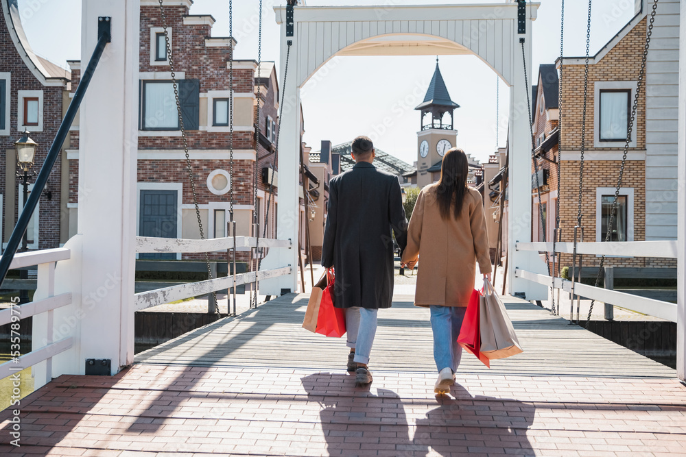 back view of interracial couple in coats holding hands and walking on bridge with shopping bags.