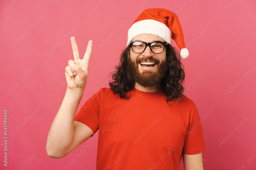 Ecstatic bearded man wearing Christmas hat is showing V gesture while smiling. Ecstatic bearded man wearing Christmas hat is showing V gesture while smiling.