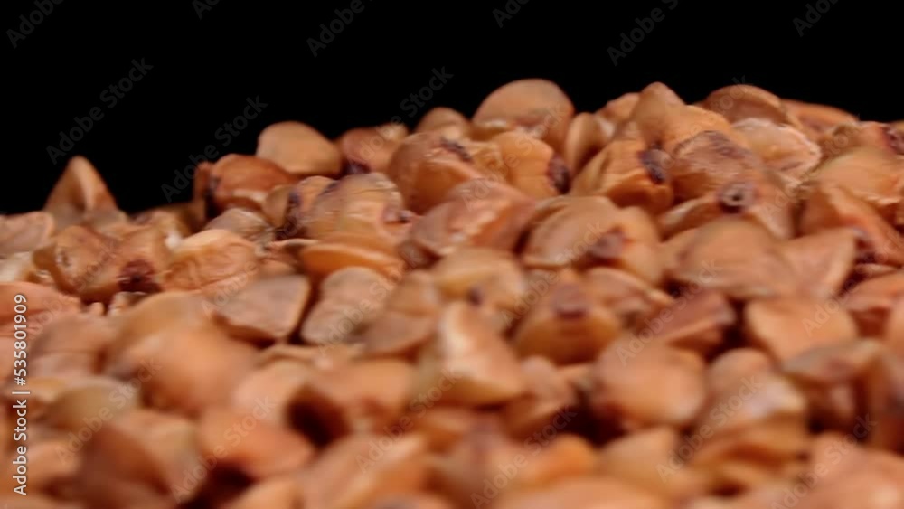 Dry Uncooked Brown Buckwheat Groats Heap on White Plate Rotating against Black Background - Macro. Raw Large Buckwheat Grains - Close-Up. Russian, Ukrainian and Belarusian Culture Dry Uncooked Brown Buckwheat Groats Heap on White Plate Rotating against Black Background - Macro. Raw Large Buckwheat Grains - Close-Up. Russian, Ukrainian and Belarusian Culture