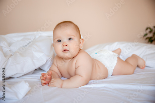 cute baby boy in a diaper is lying on a white sheet, covered with a blanket in the bedroom on the bed