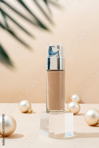A minimalistic scene of foundation bottle on glass podium with christmas decorative balls and pine tree on beige background
