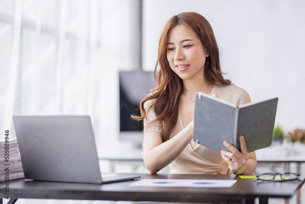 Asian Businesswoman holding notes and working on a laptop computer in the workplace, doing document planning analyzing the financial report, business plan investment, finance analysis concept. Asian Businesswoman holding notes and working on a laptop computer in the workplace, doing document planning analyzing the financial report, business plan investment, finance analysis concept.