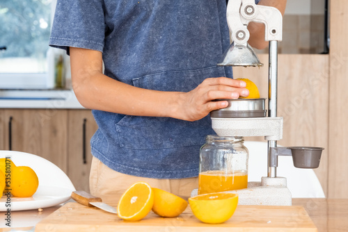 Teenager making fresh orange juice at the kitchen. Boy squeezing citrus juice with special kitchen press device.