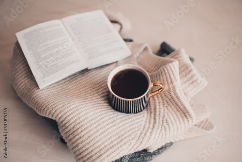 Cup of coffee stay on stack of knitted textile sweater in bed with paper open book. Winter cozy season.