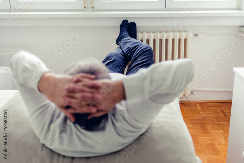 Rear view of a relaxed senior man comfortable sitting on the bed with legs on the radiator at home, looking through the large window in the winter rainy season. Man warms up feet over heater.